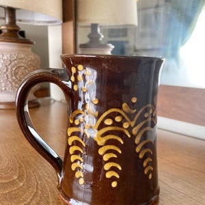 May include: A brown ceramic mug with a curved handle and a gold decorative pattern. The mug has a glossy finish and is on a wooden surface. The design features stylized leaf-like shapes.