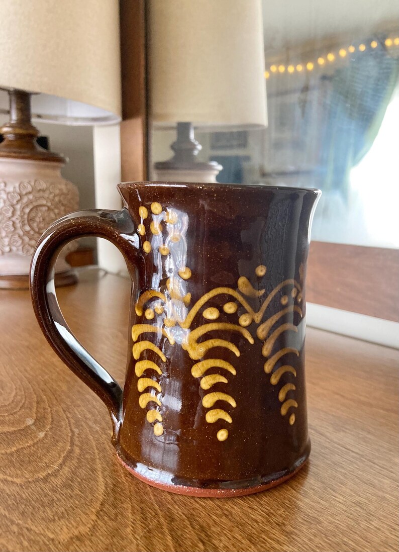 May include: A brown ceramic mug with a curved handle and a gold decorative pattern. The mug has a glossy finish and is on a wooden surface. The design features stylized leaf-like shapes.