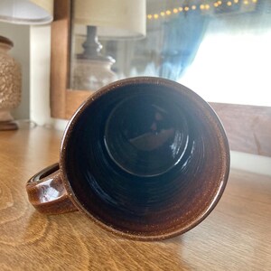 May include: A brown ceramic mug with a handle, viewed from the inside. The mug has a speckled texture and a dark interior. The mug is on a wooden surface.