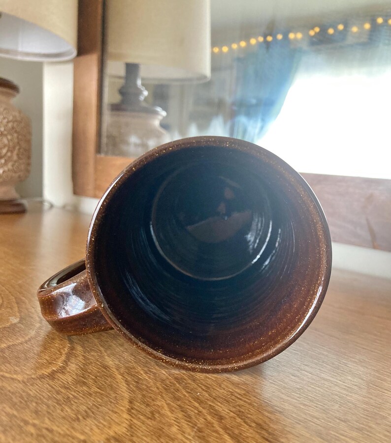 May include: A brown ceramic mug with a handle, viewed from the inside. The mug has a speckled texture and a dark interior. The mug is on a wooden surface.