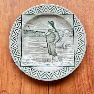 May include: A decorative plate with a green and white geometric border. The center features a monochrome illustration of a person on a beach, carrying a basket, with a ship in the background. The plate is on a wooden surface.