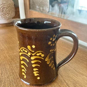 May include: A brown ceramic mug with a curved handle and a decorative yellow pattern. The mug has a glossy finish and is likely handmade. The design features curved lines and dots. Suitable for tea, coffee, or other drinks.