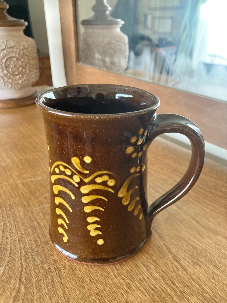 May include: A brown ceramic mug with a curved handle and a decorative yellow pattern. The mug has a glossy finish and is likely handmade. The design features curved lines and dots. Suitable for tea, coffee, or other drinks.