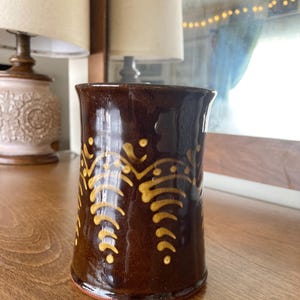 May include: A brown ceramic mug with a glossy finish. The mug features a repeating pattern of yellow, stylized leaf-like designs. The mug is cylindrical with a slightly flared rim and a red base.