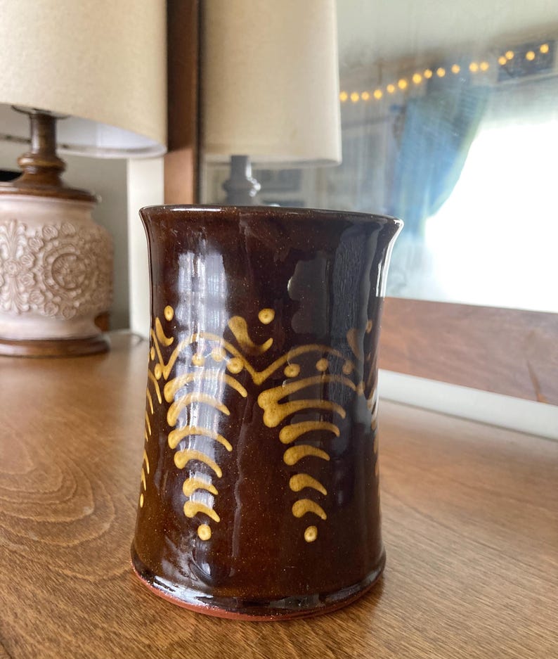 May include: A brown ceramic mug with a glossy finish. The mug features a repeating pattern of yellow, stylized leaf-like designs. The mug is cylindrical with a slightly flared rim and a red base.