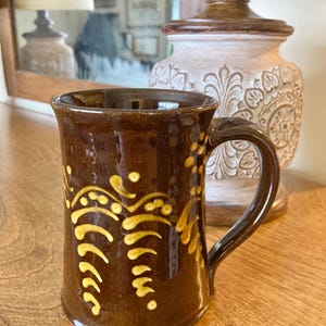 May include: A brown ceramic mug with a curved handle and a flared top. The mug features a yellow, stylized floral design. The mug is sitting on a wooden surface. A lamp and mirror are in the background.