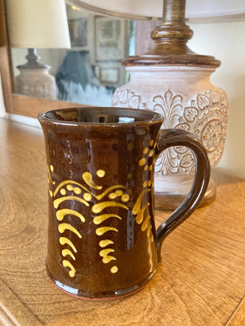 May include: A brown ceramic mug with a curved handle and a flared top. The mug features a yellow, stylized floral design. The mug is sitting on a wooden surface. A lamp and mirror are in the background.