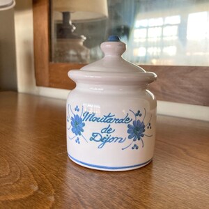 May include: A small, white ceramic jar with a lid, featuring the text "Moutarde de Dijon" in blue script, accented with blue floral designs. The jar has a light pink top and lid, and sits on a wooden surface.