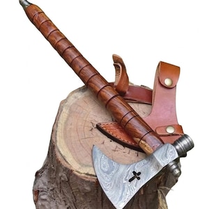 May include: A hand axe featuring a Damascus steel blade with a cross-shaped cutout and a textured metal poll. The handle is crafted from polished brown wood with segmented grooves, accompanied by a tan leather sheath with brass-colored snaps.