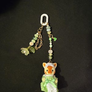 Op de afbeelding: Een sleutelhanger met een klein gevlokt dierenfiguurtje in een groen-wit geblokte jurk met een wit schort. De bedel is bevestigd aan een witte plastic ring met goudkleurige kettinkjes, lichtgroene kralen, een klein groen hartje en een bloem.