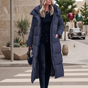 May include: A long, dark blue puffer coat with a high collar, worn over a black long-sleeve top and black trousers. The coat features a quilted design, a front zip, and side pockets. A small, dark, shiny handbag is held in one hand.