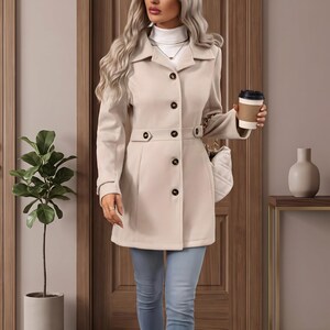 May include: A beige mid-length coat with a collar, long sleeves, and dark buttons down the front. The coat is worn over a white turtleneck top and light blue jeans, paired with beige ankle boots. A white quilted handbag is carried on the side.
