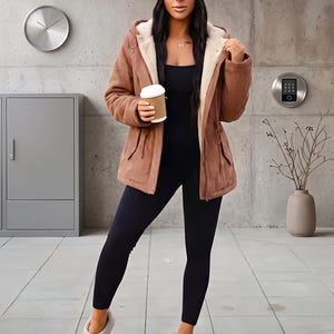 May include: A brown hooded jacket with a cream-colored faux fur lining, worn over a black bodysuit and leggings. The outfit includes a dark blue beanie and beige platform slip-on shoes. The person holds a white paper coffee cup.