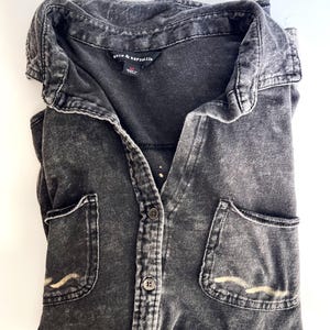 Peut inclure: Une chemise boutonnée gris anthracite avec une texture délavée à l'acide. La chemise présente un col pointu, deux poches poitrine avec une broderie ondulée beige clair et une étiquette noire au col indiquant Rock & Republic et la taille M.