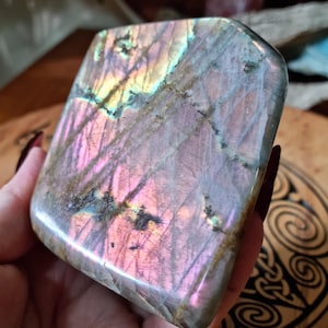 以下が含まれることがあります： 長方形の形をした、研磨されたラブラドライトの石。石の表面には、ピンク、紫、金色の虹色の輝きが見られます。石は手で持たれており、その自然な美しさとユニークな色のバリエーションを示しています。