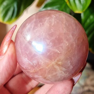 May include: A polished, spherical rose quartz crystal with a translucent, pale pink hue. The stone features natural internal fractures and cloudy inclusions that catch the light, creating a soft, reflective surface across its smooth, rounded form.