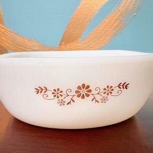 May include: A white milk glass mixing bowl featuring a brown floral pattern on the side. The bowl has a rounded shape with two small handles on opposite sides. The design consists of stylized flowers, leaves, and swirling vine accents.