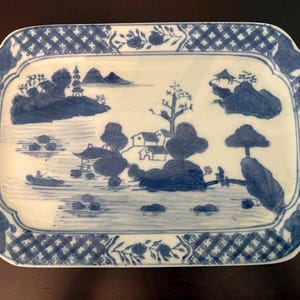 Puede incluir: Una bandeja rectangular de cerámica con un paisaje pintado a mano en azul y blanco. El centro muestra casas, árboles, montañas y un pequeño bote en el agua. El borde está decorado con un patrón de cuadrícula y motivos florales.