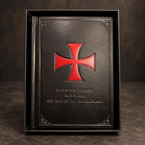May include: A black hardcover book featuring a red cross emblem centered on the front cover. Below the cross, the text reads: NON NOBIS, DOMINE, NON NOBIS, SED NOMINI TUO DA GLORIAM. The cover has decorative corner details and is presented in a black box.