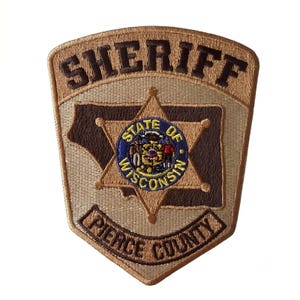 Könnte beinhalten: Ein beigefarbener und brauner bestickter Aufnäher mit dem Wort SHERIFF oben und PIERCE COUNTY unten. In der Mitte befindet sich ein sechsstrahliger Stern mit dem kreisförmigen Siegel des Staates Wisconsin in Blau, Gelb und Weiß.