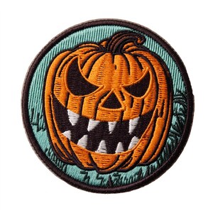 May include: A circular embroidered patch featuring an orange jack-o'-lantern with a dark brown stem, black triangular eyes, and a wide mouth filled with sharp white teeth, set against a light teal background with a dark brown stitched border.
