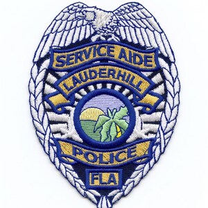 Puede incluir: Un parche bordado que presenta un águila blanca en la parte superior, un emblema circular central con un paisaje de palmera y sol, y cintas azules con el texto SERVICE AIDE, LAUDERHILL, POLICE y FLA en bordado de color oro y azul.