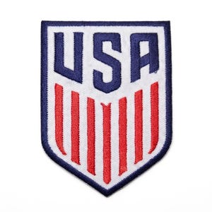 May include: A shield-shaped embroidered patch featuring the letters USA in bold dark blue thread at the top, positioned above seven vertical red stripes on a white background, all enclosed within a thick dark blue stitched border.