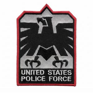 Può includere: Una toppa ricamata a forma di scudo con bordo rosso, sfondo grigio argento e un emblema di aquila nera. Il testo UNITED STATES POLICE FORCE è ricamato in lettere maiuscole bianche su una base rettangolare nera nella parte inferiore della toppa.