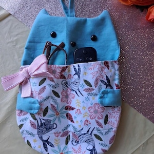 May include: A light blue fabric wall organizer shaped like a cat with a hanging loop. The front pocket features a white floral and rabbit print, a light pink ribbon bow, and two small blue fabric arms. Inside are brown glasses and a black remote.