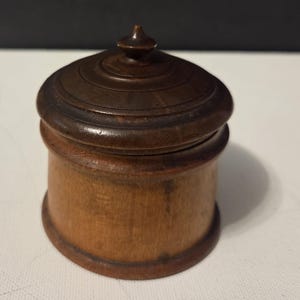 May include: A small, cylindrical wooden container with a dark brown, polished finish. It features a removable lid with a central pointed finial and concentric circular grooves. The base of the container is a lighter, natural wood tone with visible grain.
