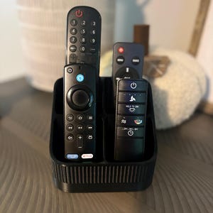 May include: A black plastic remote control organizer with vertical ribbed detailing on the front. The holder contains three black remote controls of varying sizes, featuring buttons for power, volume, navigation, and specific streaming services.