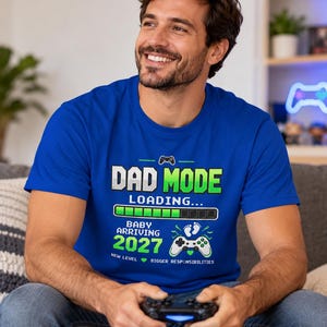 May include: A royal blue short-sleeved t-shirt featuring a graphic print with the text DAD MODE LOADING, a progress bar, BABY ARRIVING 2027, a game controller icon with baby footprints, and the text NEW LEVEL, BIGGER RESPONSIBILITIES.