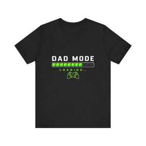 May include: A black short-sleeved t-shirt featuring the white text DAD MODE above a green and black loading bar graphic. Below the bar, the word LOADING is printed in green, followed by a green outline of a video game controller icon.