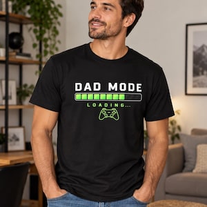 May include: A black short-sleeved t-shirt featuring the white text DAD MODE above a green loading bar graphic, the word LOADING, and a green video game controller icon centered on the chest area of the garment.