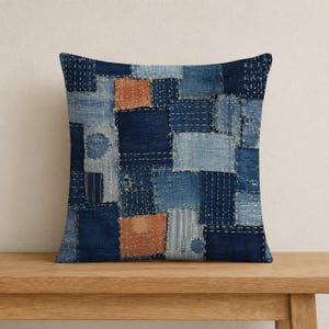 May include: A square throw pillow featuring a patchwork design of various blue and light blue fabric squares. The surface is decorated with white running stitch embroidery throughout, with two prominent orange-brown fabric patches.