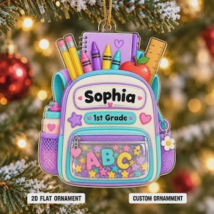May include: A flat backpack-shaped ornament featuring the name Sophia, the text 1st Grade, and ABC letters. The design includes colorful pencils, a notebook, a ruler, and an apple. The front pocket is clear with star-shaped confetti inside.