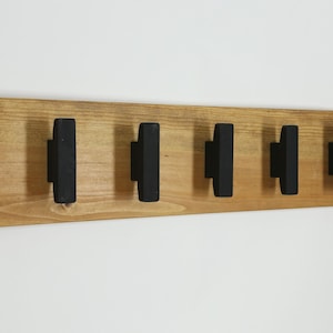 May include: A rectangular wall-mounted coat rack featuring a natural light brown wood grain base with five evenly spaced, vertical matte black hooks. The hooks are rectangular in shape and extend outward from the wooden board, providing a functional storage solution for hanging items in a home or office space.