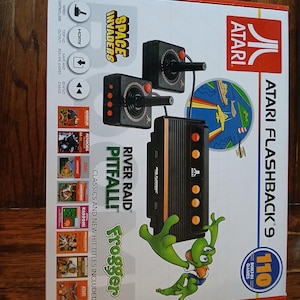 May include: Atari Flashback 9 game console box featuring 110 built-in games, two wired joysticks, and a black console unit. The packaging displays the Atari logo, game titles like Space Invaders, River Raid, Pitfall, and Frogger, and HDMI output.