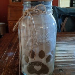 Puede incluir: Un frasco de vidrio transparente de boca ancha marca Ball con tapa de madera, atado con un lazo de cuerda marrón alrededor del cuello. Dentro del frasco hay una tela plegada a rayas blancas y beige con un gráfico de huella marrón y un corazón.