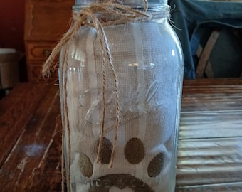Puede incluir: Un frasco de vidrio transparente de boca ancha marca Ball con tapa de madera, atado con un lazo de cuerda marrón alrededor del cuello. Dentro del frasco hay una tela plegada a rayas blancas y beige con un gráfico de huella marrón y un corazón.