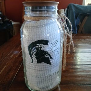 Puede incluir: Un frasco de vidrio transparente de boca ancha con tapa de madera, que contiene una toalla blanca de tejido tipo gofre con un gráfico de casco espartano verde oscuro. El cuello del frasco está envuelto con una cuerda de yute natural.
