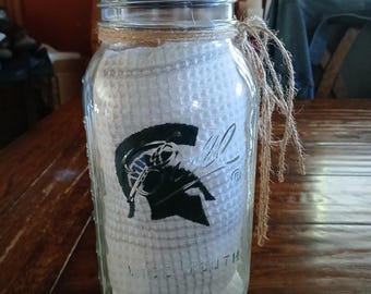 Puede incluir: Un frasco de vidrio transparente de boca ancha con tapa de madera, que contiene una toalla blanca de tejido tipo gofre con un gráfico de casco espartano verde oscuro. El cuello del frasco está envuelto con una cuerda de yute natural.