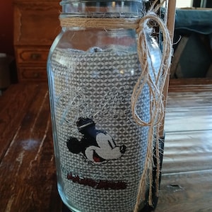 Puede incluir: Un frasco de vidrio transparente de la marca Ball con tapa de madera, relleno con una tela gris que presenta un patrón geométrico blanco repetitivo y un gráfico de Mickey Mouse. El frasco tiene un lazo de yute natural atado al cuello.