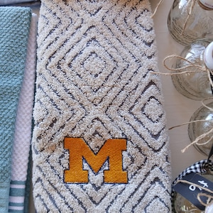 Puede incluir: Una toalla de mano rectangular de felpa con un patrón geométrico de diamantes en color crema y azul oscuro. La toalla está bordada con una letra M grande y llamativa en amarillo dorado con un contorno azul oscuro cerca del borde inferior.