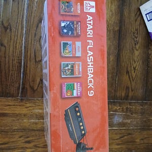 Puede incluir: Una caja de consola de juegos Atari Flashback 9 de color naranja con el logotipo y el texto de Atari. La caja muestra t&iacute;tulos de juegos como Adventure, Asteroids, Centipede, Jungle Hunt y Kaboom, junto a una ilustraci&oacute;n de la consola y dos joysticks.