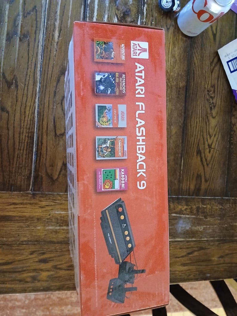 Puede incluir: Una caja de consola de juegos Atari Flashback 9 de color naranja con el logotipo y el texto de Atari. La caja muestra t&iacute;tulos de juegos como Adventure, Asteroids, Centipede, Jungle Hunt y Kaboom, junto a una ilustraci&oacute;n de la consola y dos joysticks.