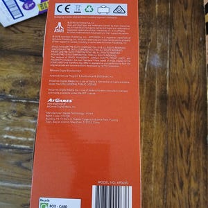 Puede incluir: Una caja de producto rectangular de color naranja con el texto Age 15+, Atari, AtGames y varios avisos de derechos de autor. La caja incluye un c&oacute;digo de barras con el n&uacute;mero 8 18858 02963 6, un s&iacute;mbolo de reciclaje y el modelo AR3050.