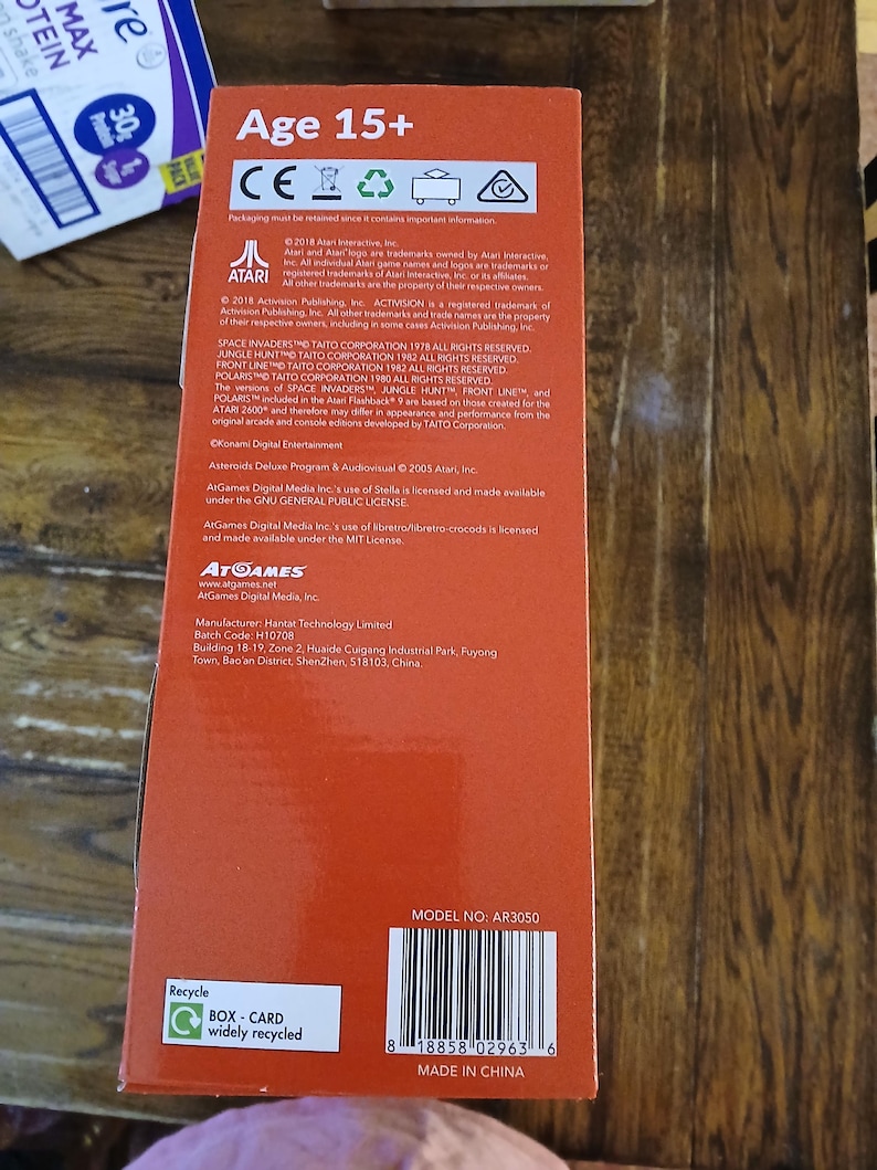 Puede incluir: Una caja de producto rectangular de color naranja con el texto Age 15+, Atari, AtGames y varios avisos de derechos de autor. La caja incluye un c&oacute;digo de barras con el n&uacute;mero 8 18858 02963 6, un s&iacute;mbolo de reciclaje y el modelo AR3050.