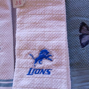Puede incluir: Un paño de cocina blanco texturizado con un logotipo de león azul bordado y la palabra LIONS en letras azules. El paño tiene un patrón de tejido de gofre e incluye una pequeña etiqueta blanca unida a la esquina superior con el texto 8$.