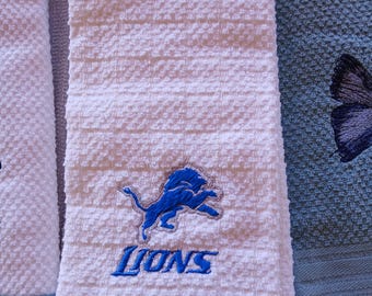 Puede incluir: Un paño de cocina blanco texturizado con un logotipo de león azul bordado y la palabra LIONS en letras azules. El paño tiene un patrón de tejido de gofre e incluye una pequeña etiqueta blanca unida a la esquina superior con el texto 8$.